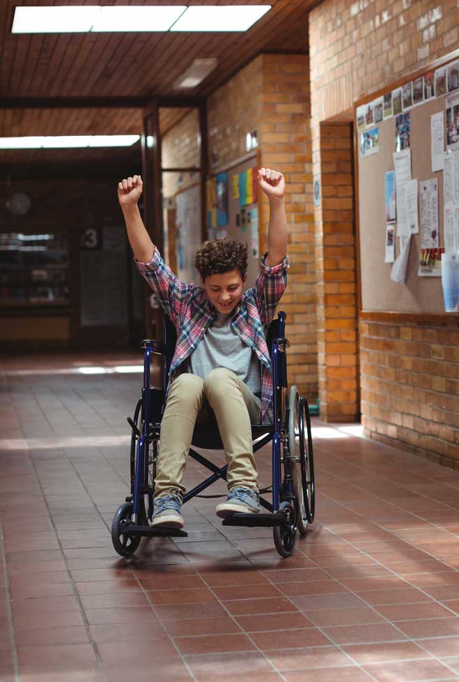 student in wheelchair in school hallway. His arms and hands are held above him, triumphantly.